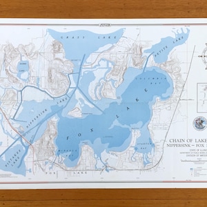 Peut inclure: Une carte vintage de la région des lacs Chain, comprenant les lacs Nippersink et Fox, dans l'Illinois. La carte présente des plans d'eau bleus, des contours terrestres beiges et une rose des vents. Le titre "Chain of Lakes Region" est visible.