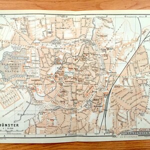 May include: A vintage map of Münster, Germany, printed in black and white with orange highlights. The map shows the city's streets, parks, and landmarks, including the "Schloss Garten" and the "Zentral Friedhof". The map's title is "Münster 1:12,500" and includes a scale bar.