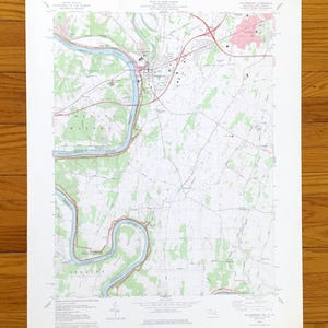 Puede incluir: Un mapa topográfico antiguo de una zona con un río, carreteras y edificios. El mapa presenta tonos de verde, azul y rojo, con líneas de contorno y nombres de lugares. El mapa está sobre papel blanco y sobre una superficie de madera.