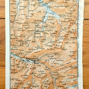 May include: A vintage map of the Swiss Alps, showing the area around the Muotatal valley. The map is printed in brown, orange, and blue ink on a white background. The map includes the names of mountains, rivers, and towns. The map is titled "Kilometer 1:150.000".