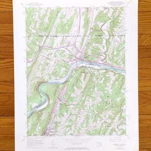 Puede incluir: Un mapa topográfico antiguo de un valle fluvial, con tonos verdes, azules y morados. El mapa, impreso en papel blanco, muestra curvas de nivel y características geográficas. El mapa está sobre una superficie de madera.