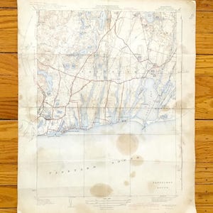 Può includere: Mappa vintage di Vineyard Sound e Falmouth, Massachusetts, con tonalità blu e marrone sbiadite. La mappa mostra dettagliate caratteristiche geografiche, tra cui coste, strade e corpi idrici. La carta presenta pieghe e macchie, che ne esaltano il fascino antico.