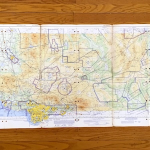 May include: A vintage aviation sectional chart, unfolded and laid flat on a wooden surface. The map features detailed lines, symbols, and color-coded areas, including yellow, green, and blue, representing geographical features and airspace information.