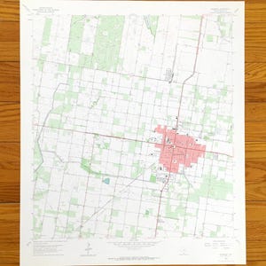 Peut inclure: Une carte topographique détaillée avec un quadrillage de routes, des zones ombragées en vert et une zone urbaine centrale en rose. La carte est imprimée sur papier blanc et comporte du texte en haut et en bas. La carte est posée sur une surface en bois.