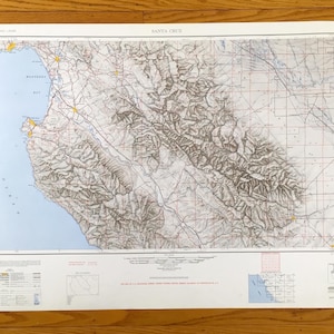 May include: A detailed map of Santa Cruz, California, featuring coastal areas, mountains, and roads. The map has a light blue ocean and a white border. The map includes the text "SANTA CRUZ" at the top.