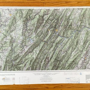 Puede incluir: Un mapa topográfico detallado del área de Cumberland, con curvas de nivel, ríos y carreteras. El mapa está impreso en una hoja de papel grande y es principalmente verde y blanco, con acentos amarillos. El mapa se muestra sobre una superficie de madera.