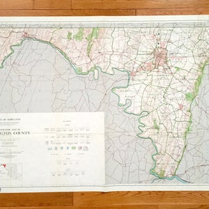 Puede incluir: Un mapa topográfico antiguo del condado de Washington, Maryland, con detalles geográficos. El mapa presenta una paleta de colores verde, rosa y beige, con texto que indica el título y la escala. El mapa está desplegado sobre una superficie de madera.