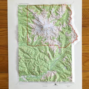 May include: A topographic map of a mountainous region, featuring shades of green, white, and brown. The map is framed in white and has a red border outlining a specific area. The map appears to be a 3D rendering of the terrain.