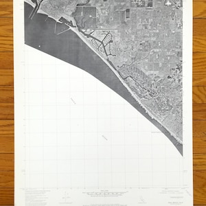Può includere: Fotografia aerea in bianco e nero di una città costiera. L'immagine mostra un grande specchio d'acqua, una costa e una città con strade ed edifici. Il testo "PACIFIC OCEAN" è visibile nell'angolo in basso a sinistra dell'immagine.