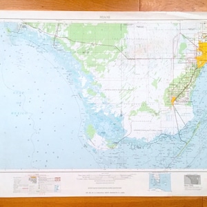 May include: A vintage map of Miami, Florida, showing the city's layout and surrounding areas. The map is printed on paper and has a light blue background with yellow and green markings. The map includes the text "MIAMI" in the top center and "GULF OF MEXICO" in the bottom left corner.