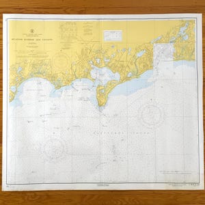 Può includere: Una carta nautica intitolata "Hyannis Harbor and Vicinity" con dettagli di elementi terrestri e acquatici. La mappa utilizza colori giallo, blu e bianco, con linee di griglia ed etichette di testo. La carta è su carta bianca, su una superficie di legno.
