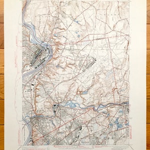 Puede incluir: Un mapa topográfico antiguo de una zona de Massachusetts, con líneas de contorno detalladas, carreteras y elementos acuáticos. El mapa está impreso en tonos de marrón, azul y gris, con texto que indica el origen y la escala del mapa. El mapa se muestra sobre una superficie de madera.