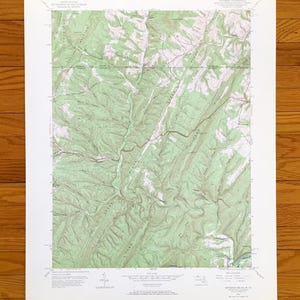 Puede incluir: Un mapa topográfico detallado de una región montañosa, con curvas de nivel e indicaciones de altitud. El mapa, impreso en papel blanco, incluye texto que indica su origen y escala. El mapa está sobre una superficie de madera.