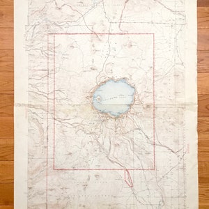 May include: Vintage topographic map featuring Crater Lake, Oregon, with detailed contour lines, water features, and place names. The map has a red border and is printed on aged paper, showing signs of wear and tear. The map is folded and laid out on a wooden surface.