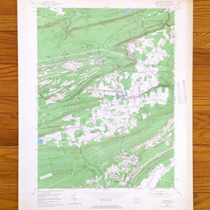Puede incluir: Un mapa topográfico vintage con detalles de terreno verdes y blancos, incluyendo curvas de nivel, carreteras y elementos acuáticos. El mapa está impreso en papel envejecido, con texto en la parte superior e inferior.