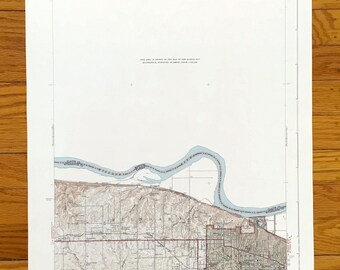 Puede incluir: Un mapa topográfico antiguo de una ciudad, con líneas de contorno detalladas, carreteras y edificios. El mapa incluye un río azul y texto que indica la escala y el área geográfica. El mapa está impreso en papel envejecido.