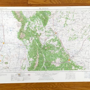 May include: A detailed map of Trinidad, showing green shaded areas representing mountainous regions and a network of roads and towns. The map is printed on a white sheet of paper and is displayed on a wooden surface.