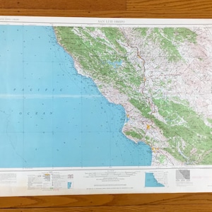 May include: A vintage map of San Luis Obispo, California, showing the coastline, mountains, and roads. The map is printed on paper and has a blue and green color scheme. The text "PACIFIC OCEAN" is printed in the lower left corner of the map.