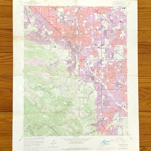 May include: A vintage topographic map with detailed color-coded areas. The map features shades of green, pink, purple, and white, depicting terrain and urban areas. The map is creased and shows signs of age, resting on a wooden surface.