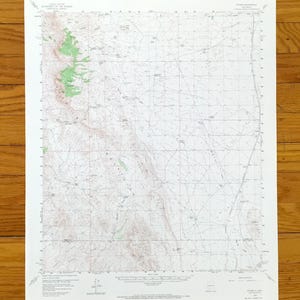 May include: A vintage topographic map with a grid pattern, featuring green shaded areas and brown contour lines. The map is printed on white paper and is set against a wooden background.