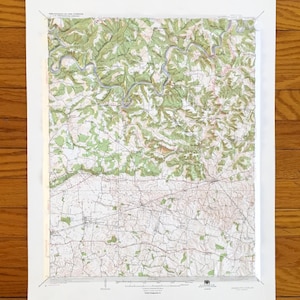 Może przedstawiać: Oprawiona mapa topograficzna z białym obramowaniem. Mapa przedstawia zielone i brązowe cieniowanie, z niebieskimi liniami reprezentującymi rzeki. Mapa jest zamontowana na białej powierzchni i jest wyświetlana na drewnianej podłodze.