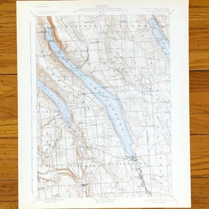 May include: A vintage map print of the Finger Lakes region in New York State. The map features detailed topographical lines and the names of the lakes and surrounding towns. The lakes are colored in light blue, contrasting with the beige and brown land features.