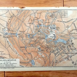 May include: A vintage map of Wilhelmshöhe, Germany, showing the surrounding area, including the Wilhelmshöhe Palace, the Berg Park, and the surrounding town of Kassel. The map is printed in brown, blue, and black ink on a white background. The map includes a scale bar and a legend.