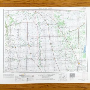 以下が含まれることがあります： スリーリバーフォールズの詳細な地図。道路と地理的特徴を示すグリッドパターン。地図には上下にテキストがあり、白い境界線と木製の背景があります。