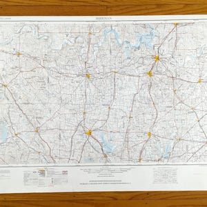 May include: A detailed map of Sherman, featuring roads, rivers, and towns. The map is printed on a white sheet with a grid pattern and various colors. The title "SHERMAN" is visible at the top. The map is lying on a wooden surface.
