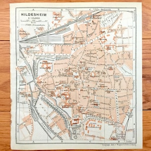 May include: A vintage map of Hildesheim, Germany, printed in black and white with red and orange highlights. The map shows the city's streets, buildings, and landmarks, including the Hauptbahnhof (main train station), the Andreas Kirche (Andreas Church), and the Stadtkirche (city church). The map is dated 1900 and is in good condition.