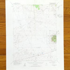May include: A vintage topographic map with a grid pattern, featuring contour lines and shaded green areas. The map is printed on white paper and shows geographical features. The map is lying on a wooden surface.