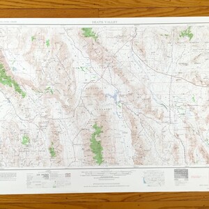 May include: A detailed topographic map of Death Valley, featuring contour lines, elevation markings, and geographical features. The map includes the text "Death Valley" at the top and is printed on white paper. The map is displayed on a wooden surface.