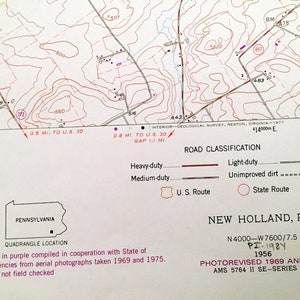 Op de afbeelding: Een zwart-witte kaart van New Holland, Pennsylvania, met een paarse omtrek van de staat. De kaart toont wegen, hoogte en andere geografische kenmerken. De kaart is getiteld "New Holland, PA." en bevat de datum "PI-1984 1956".