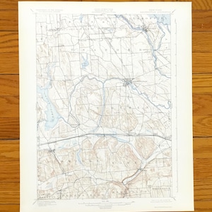 May include: A vintage topographical map of the Baldwinsville, New York area. The map features detailed contour lines, roads, and water features in blue and brown hues. The map is printed on aged paper and is set against a wooden background.