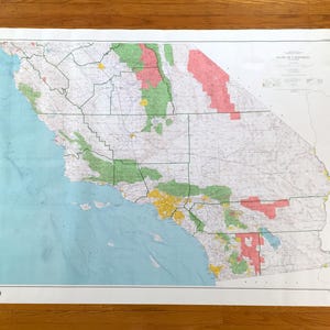 May include: A large, detailed map of California, featuring a variety of colors to denote different regions. The map is printed on a white sheet of paper and is displayed on a wooden surface. The map includes the state's coastline and major cities.