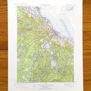 Puede incluir: Un mapa topográfico de Plymouth, Massachusetts, impreso en papel blanco. El mapa presenta áreas forestales verdes, cuerpos de agua azules y una red de líneas marrones y rojas que representan carreteras e infraestructura costera.