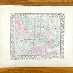 Könnte beinhalten: Eine antike Karte mit dem Titel "PLAN of BALTIMORE" mit einem dekorativen Rahmen. Die Karte zeigt eine detaillierte Darstellung der Stadt mit Straßen und nummerierten Bezirken in Rosa, Grün und Blau. Das Papier hat ein leicht gealtertes Aussehen.