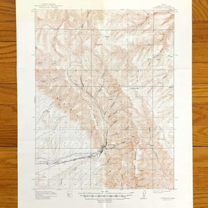 Op de afbeelding: Een vintage topografische kaart met een rasterpatroon en contourlijnen, die een bergachtig gebied weergeeft. De kaart is gedrukt in bruin- en tinten, met tekst bovenaan die de uitgevende instantie aangeeft. De kaart is op wit papier.