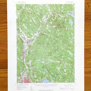 May include: A detailed topographic map of a rural area, featuring green and brown terrain, blue lakes, and a river. The map is printed on white paper and includes text and symbols indicating geographical features. The map is folded and laid on a wooden surface.