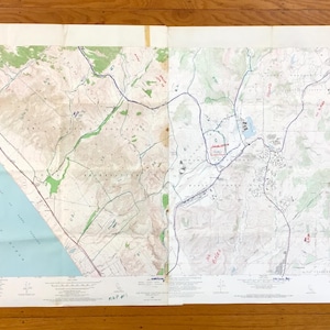 Pode incluir: Um mapa topográfico vintage, dobrado e colocado sobre uma superfície de madeira. O mapa apresenta linhas de contorno detalhadas, cursos de água azuis e áreas sombreadas em verde, com notas manuscritas.