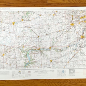 May include: A vintage map titled "Aurora" with detailed road and geographical features. The map features a grid pattern with various colors, including red, yellow, and green, on a white background. The map is displayed on a wooden surface.