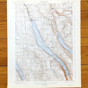 May include: A vintage topographic map of New York State, featuring blue lakes and brown shaded relief. The map shows the area's elevation and geographical features, with place names and grid lines. The map is printed on aged paper.