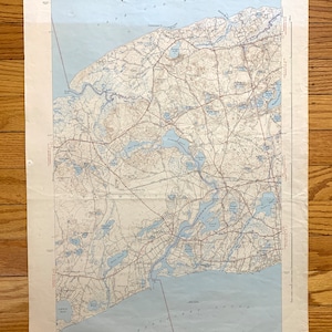 Può includere: Una mappa vintage della zona di Plymouth, Massachusetts. La mappa mostra strade, fiumi, laghi e la costa. La mappa è stampata su carta color crema con linee blu e rosse. La mappa è intitolata "Stato del Massachusetts, Dipartimento dell'Interno, Indagine Geologica, Quadrante di Plymouth, Massachusetts, 1948."