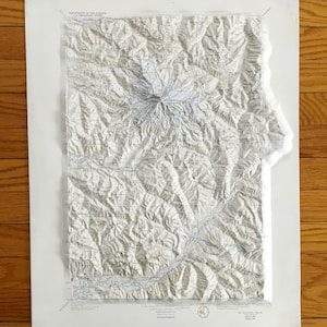 May include: A topographic map of Mount Rainier, with detailed relief shading, mounted on a white backing. The map features blue lines representing rivers and the text "MT RAINIER". The map is on a wooden surface.