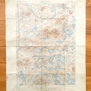 May include: A vintage map of the Adirondack Mountains in New York State, showing the location of lakes, rivers, and mountains. The map is printed on a cream-colored paper and has a blue and brown color scheme.