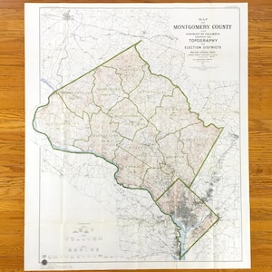 May include: A vintage map of Montgomery County, Maryland, showing the District of Columbia, topography, and election districts. The map features a detailed layout with green borders and text, printed on aged paper. The map is folded and displayed on a wooden surface.