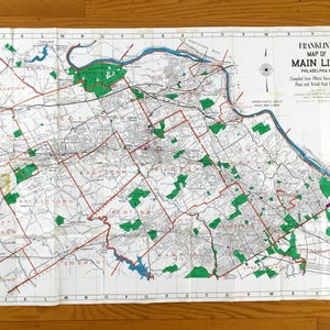 Könnte beinhalten: Eine alte Papierkarte mit dem Titel Franklin's Map of Main Line Philadelphia, PA. Die Karte zeigt ein Gitternetz mit Buchstaben und Zahlen, schwarze Linien für Straßen und Städte, blaue Wasserflächen und grüne Bereiche für Parkanlagen.