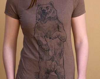 Könnte beinhalten: Ein braunes T-Shirt mit einer Schwarz-Weiß-Illustration eines Bären, der auf seinen Hinterbeinen steht. Der Bär ist in einer einfachen, stilisierten Art und Weise gezeichnet. Der Text "Weatherly" ist unter dem Bären gedruckt.