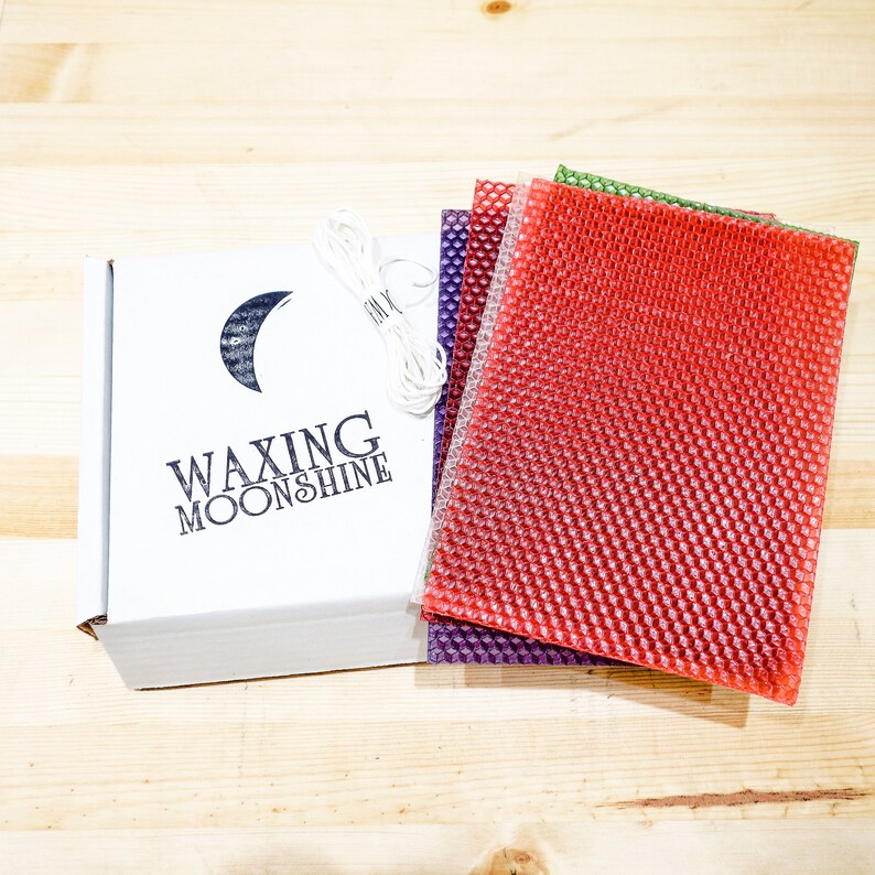 May include: A white box with the text "Waxing Moonshine" and a crescent moon logo, alongside sheets of red, purple, and green honeycomb wax. A bundle of white wick is also included. The items are arranged on a light wood surface.