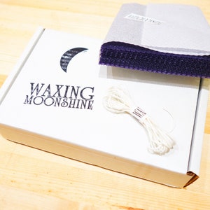 May include: A white box with the text "WAXING MOONSHINE" and a crescent moon logo. The box contains purple sheets, and a bundle of white string. The items are on a light wood surface.